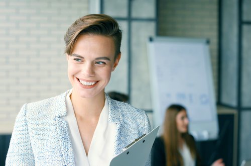 A smiling woman in a blue blazer is holding a clipboard while standing in a modern office. In the background, other people can be seen.