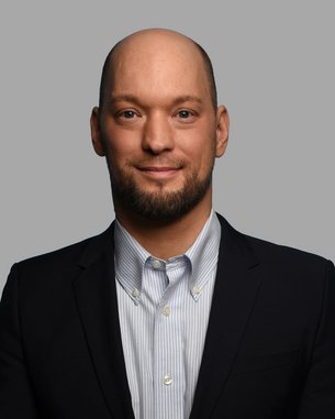 A man with a bald head and a beard is wearing a black suit and a light blue shirt. He is looking directly at the camera.