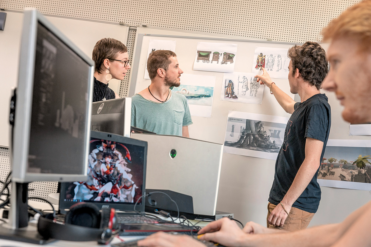 Vier Personen stehen um eine Wand mit Konzeptzeichnungen. Einer zeigt auf die Zeichnungen, während die anderen aufmerksam zuhören. Im Vordergrund sind Computer und Kopfhörer sichtbar.