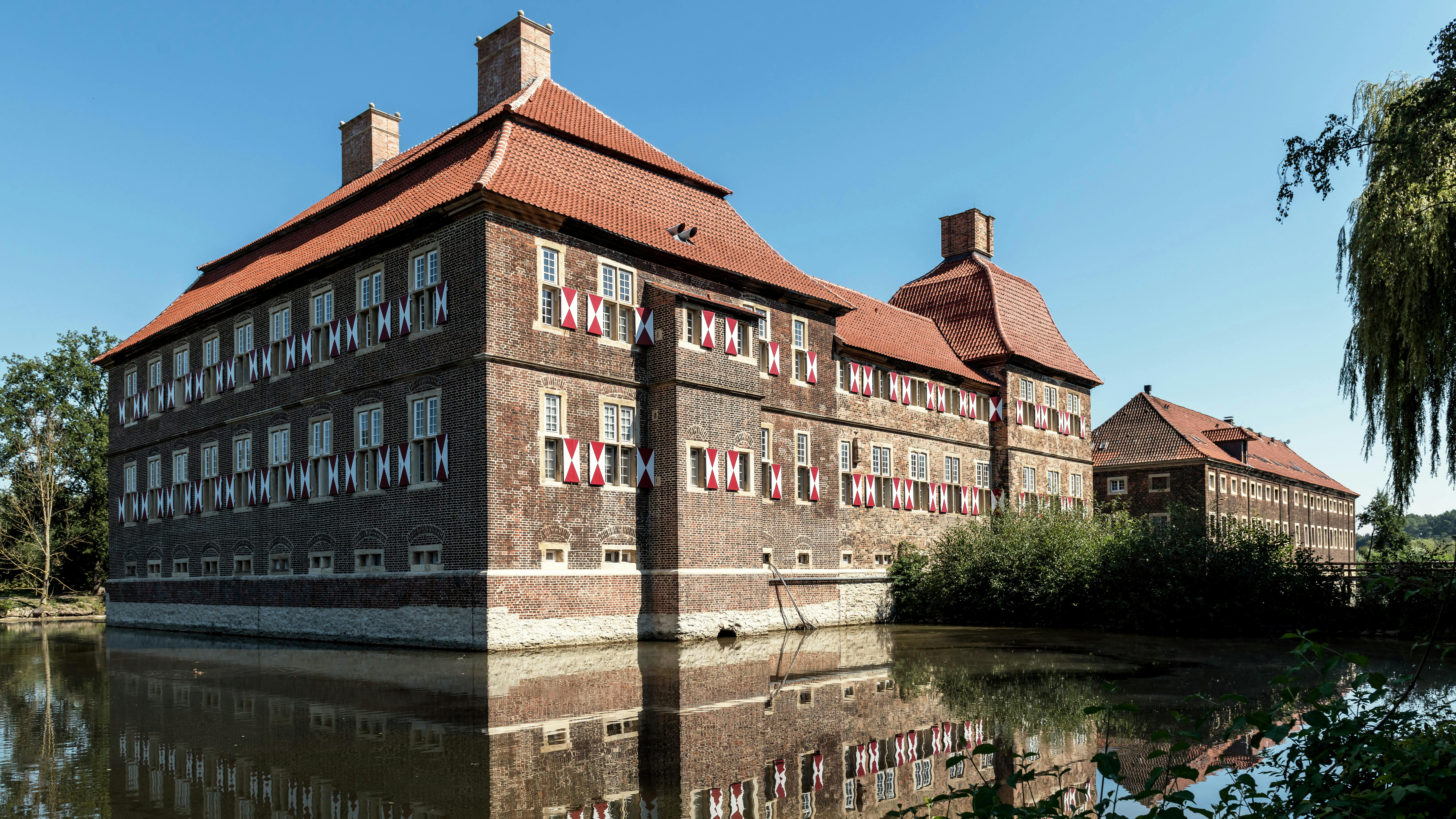 Historic building with a red gabled roof and windows adorned with colorful curtains, surrounded by water and trees. The architecture showcases a blend of brick and modern elements.