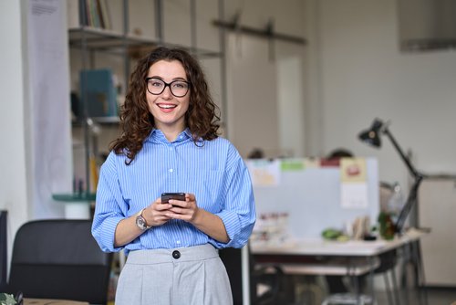 A woman with curly hair and glasses stands in a modern office, holding a smartphone in her hand. She is wearing a striped shirt.