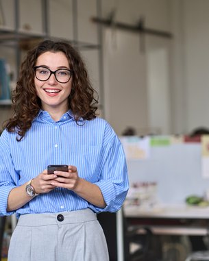 A woman with curly hair and glasses stands in a modern office, holding a smartphone in her hand. She is wearing a striped shirt.