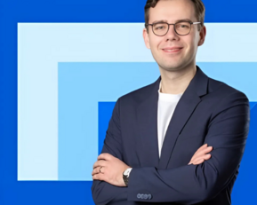 A young man with glasses stands smiling in front of a blue background. He is wearing a black blazer and has his arms crossed.