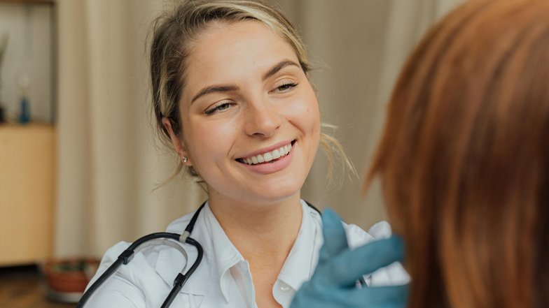 Eine lächelnde Ärztin untersucht das Haar einer Patientin. Die Ärztin trägt Handschuhe und hat ein Stethoskop um den Hals. Im Hintergrund sind einige Möbel und Pflanzen sichtbar.