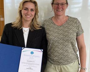 Zwei Frauen stehen nebeneinander, eine hält ein Zertifikat für den Wissenschaftspreis 2025 in der Hand. Beide lächeln in die Kamera.