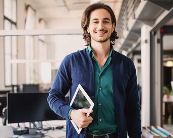Ein junger Mann mit langen, lockigen Haaren trägt einen blauen Cardigan und ein grünes Hemd. Er hält ein Tablet in der Hand und steht in einem modernen Büro mit Schreibtischen und Computern im Hintergrund.