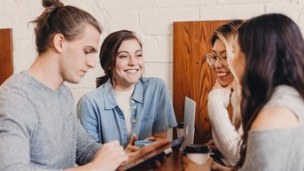 Four people are sitting at a table in a café. A man is using a tablet while the other three women are smiling and chatting with each other. Drinks are placed on the table, and the atmosphere feels relaxed.