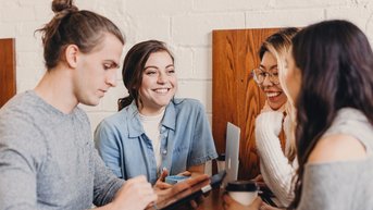 Four people are sitting at a table in a café. A man is using a tablet while the other three women are smiling and chatting with each other. Drinks are placed on the table, and the atmosphere feels relaxed.