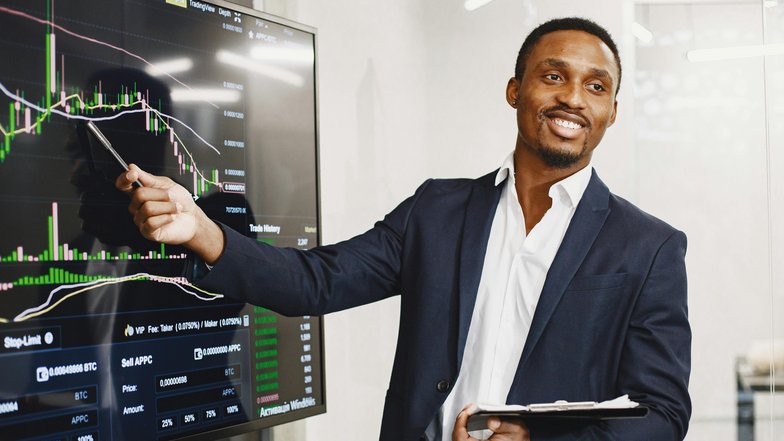 Ein Mann in einem Anzug zeigt auf ein Finanzdiagramm auf einem Bildschirm und erklärt die Daten. Er hält ein Notizbuch in der Hand und lächelt, während er seine Präsentation durchführt.