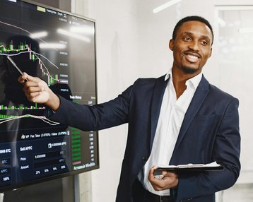 A man in a suit points to a financial chart on a screen and explains the data. He holds a notebook in his hand and smiles as he delivers his presentation.