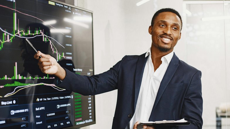 A man in a suit points to a financial chart on a screen and explains the data. He holds a notebook in his hand and smiles as he delivers his presentation.