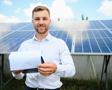 Mann in Hemd hält ein Dokument vor Solarpanelen und lächelt. Die Umgebung zeigt eine sonnige Landschaft mit blauen Himmel.