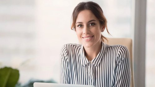 Eine Frau mit langen, braunen Haaren sitzt an einem Schreibtisch und lächelt. Sie trägt ein gestreiftes Hemd und hat einen Laptop vor sich. Im Hintergrund sind unscharfe Pflanzen und Fenster zu sehen.