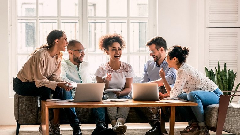 Fünf Personen sitzen um einen Tisch mit Laptops, lachen und diskutieren. Sie scheinen an einem kreativen Projekt oder einer Besprechung zu arbeiten. Der Raum hat große Fenster und eine helle, einladende Atmosphäre.