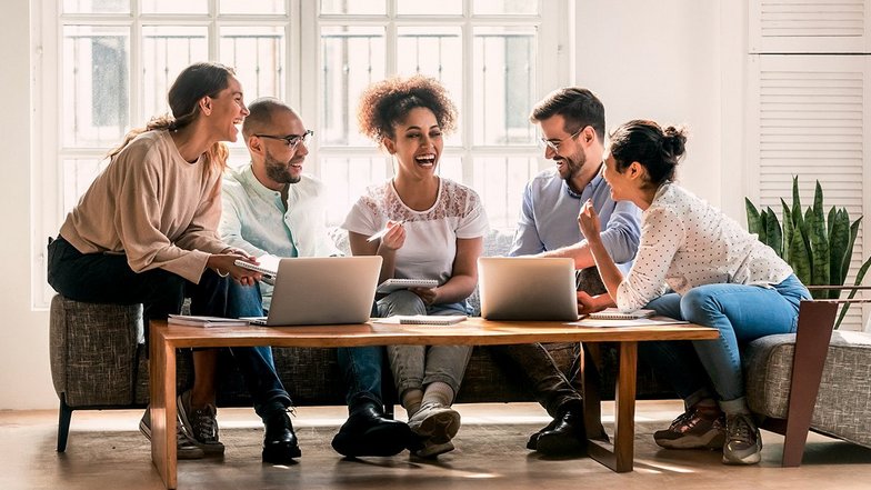 Five people are sitting around a table with laptops, laughing and discussing. They seem to be working on a creative project or having a meeting. The room has large windows and a bright, inviting atmosphere.
