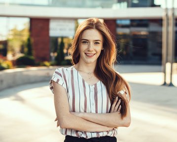 Junge Frau mit langen, lockigen Haaren und gestreiftem Oberteil, die lächelnd mit verschränkten Armen in einem urbanen Umfeld steht.