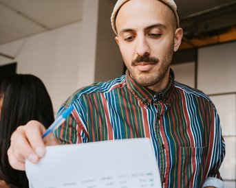 A man in a striped shirt and a cap is looking at a piece of paper while holding a pen. On the table, writing materials and books are visible. In the background, another person can be seen.