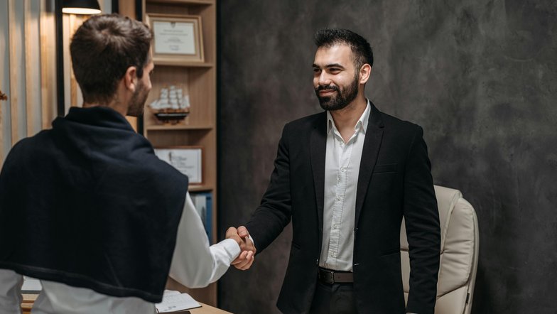 Zwei Männer schütteln sich die Hände in einem Büro. Der Mann im Anzug lächelt, während der andere, leger gekleidet, ihm gegenübersteht. Auf dem Tisch liegen Dokumente und ein Stuhl ist im Hintergrund sichtbar.