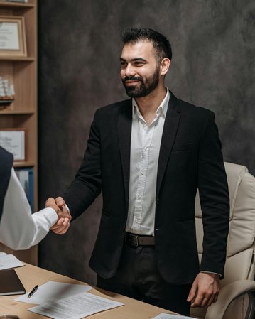 Two men are shaking hands in an office. The man in the suit is smiling, while the other, dressed casually, stands across from him. Documents are spread out on the table, and a chair is visible in the background.