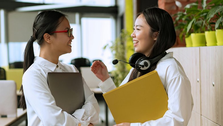 Kategorie Kommunikation SRH University Two women are smiling as they chat in a modern office. One is holding a laptop, while the other has headphones and a yellow folder in her hand. Plants and office furniture are visible in the background.