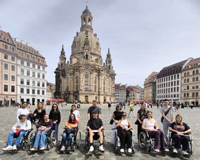 Gruppierung von Menschen in Rollstühlen auf einem Platz vor einer historischen Kirche, umgeben von Gebäuden im Hintergrund.
