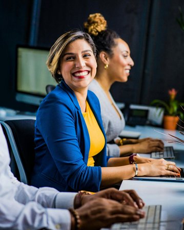Three people are sitting at a table in a modern office. A woman is smiling warmly at the camera, while the others are focused on their laptops. Plants are arranged on the table.
