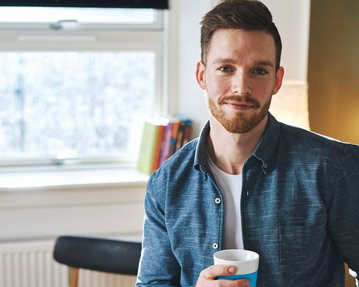 Ein Mann mit Bart hält eine Tasse in der Hand und lächelt. Er trägt ein blaues Hemd und steht in einem hellen Raum mit Pflanzen im Hintergrund.