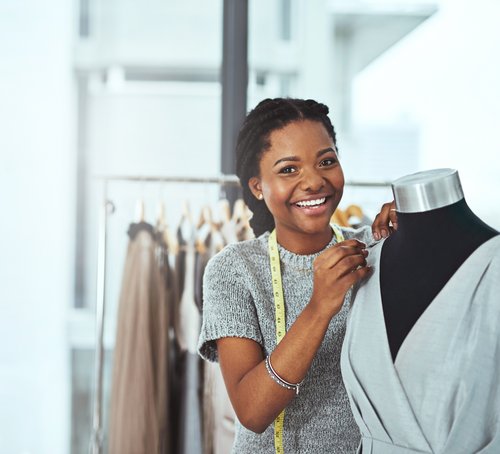 A smiling woman with a braid stands next to a dress form, adjusting a gray dress. In the background, clothes are hanging on racks.