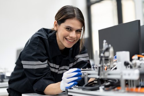 A young woman in work attire is operating a machine and holding a tool in her hand while smiling.