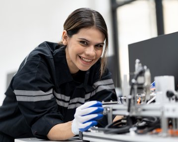 A young woman in work attire is operating a machine and holding a tool in her hand while smiling.