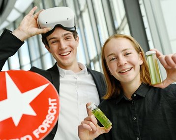 Zwei junge Menschen lächeln, einer trägt eine VR-Brille und hält ein Schild mit dem Text "Hier forscht". Die andere Person zeigt zwei Fläschchen.
