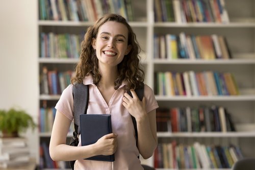 Eine junge Frau mit lockigem Haar trägt einen Rucksack und hält ein Buch in der Hand, lächelnd vor einem Bücherregal.