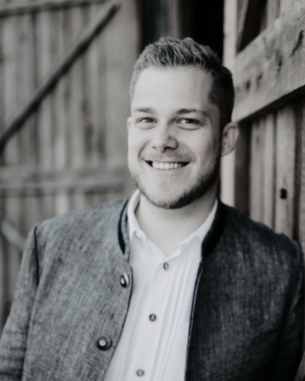 A smiling man in traditional clothing stands next to a wooden door. His face is clearly visible, with short hair and a well-groomed beard.