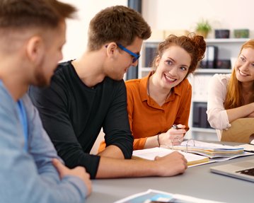 Vier junge Erwachsene sitzen an einem Tisch und diskutieren. Eine Frau lächelt, während die anderen aufmerksam zuhören. Bücher und Notizen liegen auf dem Tisch.