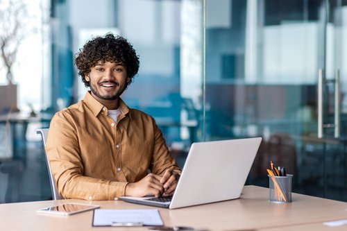 A young man with curly hair sits at a desk, smiling as he works on a laptop. Writing utensils are visible.
