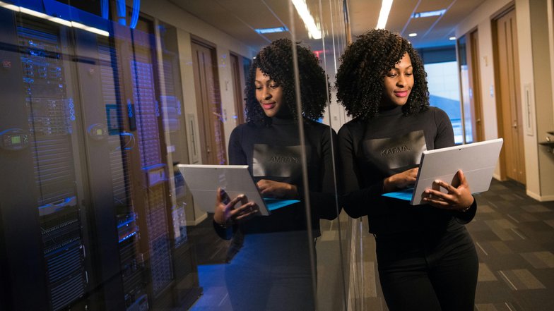Kategorie Data Science studieren Studium SRH University A woman with curly hair stands in a modern office, looking at a tablet. She is wearing a black top and is positioned in front of a reflective wall. In the background, servers are visible.