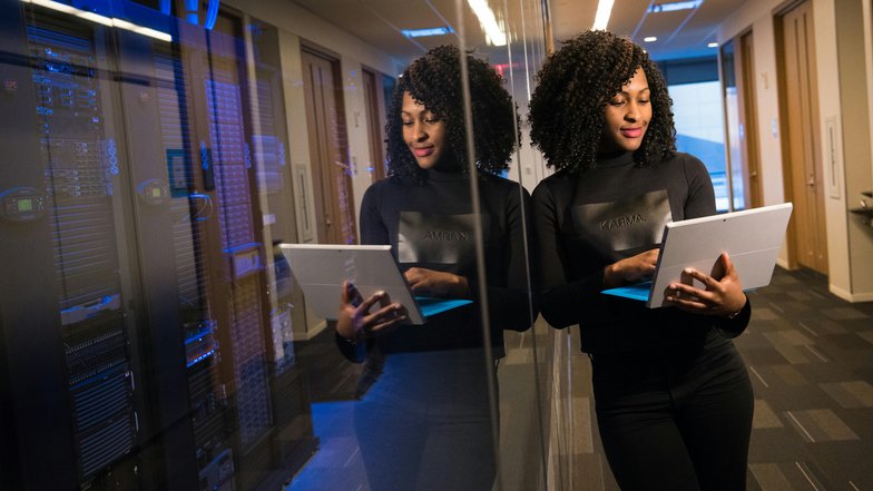 Kategorie Data Science studieren Studium SRH University A woman with curly hair stands in a modern office, looking at a tablet. She is wearing a black top and is positioned in front of a reflective wall. In the background, servers are visible.
