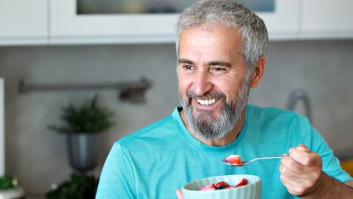 An older man with a gray beard smiles as he eats fruit from a bowl with a spoon.
