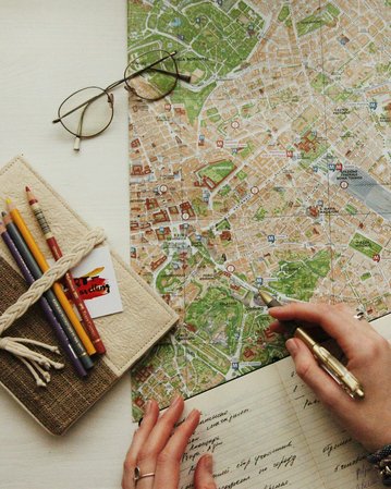 Eine Person hält einen Stift und zeigt auf eine Stadtkarte, die auf einem Tisch liegt. Neben der Karte befinden sich eine Notizbuch und bunte Stifte. Eine Brille liegt ebenfalls auf dem Tisch.