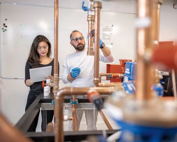 Eine Frau mit Brille hält ein Blatt Papier, während ein Mann mit Bart und Schutzbrille an einer Kupferrohrinstallation arbeitet. Im Hintergrund sind technische Geräte und eine Tafel sichtbar.