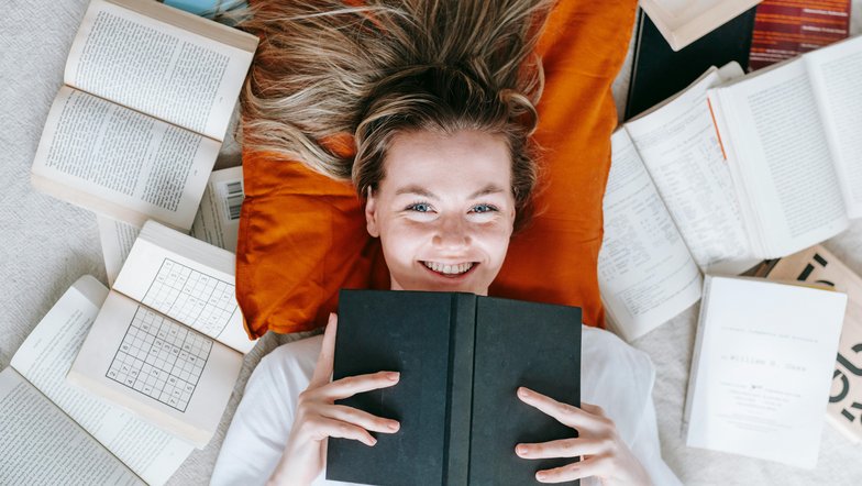 Kategorie Psychologie studieren Studium SRH University A smiling woman is lying on an orange pillow, surrounded by open and closed books. She holds a black book in front of her and looks directly at the camera.
