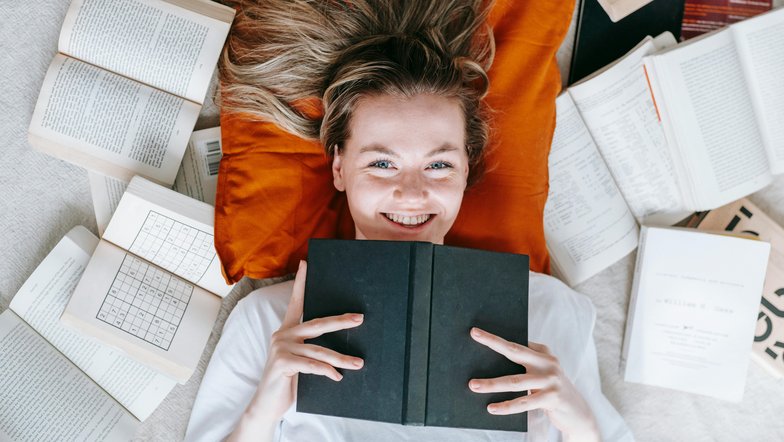 Kategorie Psychologie studieren Studium SRH University Eine lächelnde Frau liegt auf einem orangefarbenen Kissen, umgeben von offenen und geschlossenen Büchern. Sie hält ein schwarzes Buch vor sich und schaut direkt in die Kamera.