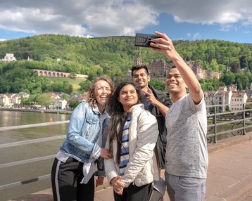 Studierende auf der Alten Brücke in Heidelberg, welche gerade ein Selfie mit Blick auf das Heidelberger Schloss schießen.