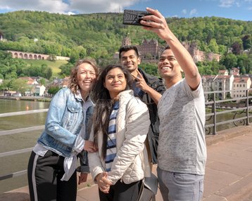 Studierende auf der Alten Brücke in Heidelberg, welche gerade ein Selfie mit Blick auf das Heidelberger Schloss schießen.