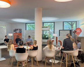 Eine Gruppe von Personen sitzt in einem modernen Raum und diskutiert. An den Wänden sind Flipcharts und Präsentationsmaterialien sichtbar.