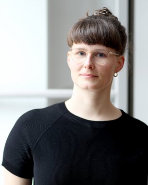 Eine Person mit kurzen, braunen Haaren und Brille trägt ein schwarzes Oberteil und schaut in die Kamera.