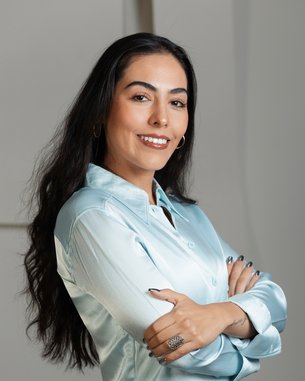 A woman with long, dark hair is wearing a light blue shirt and smiling as she poses with her arms crossed.
