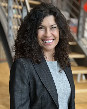 A woman with curly brown hair is wearing a gray sweater and a black blazer. She is smiling and standing in front of a staircase.