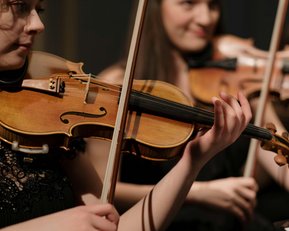 Two female musicians are playing violins in an orchestra. The first musician holds her violin with one hand and plays, while the second musician in the background is also playing a violin. Both are dressed in black.
