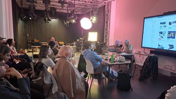 A group of people is sitting in a room, listening to a presentation while a screen displays technical information.